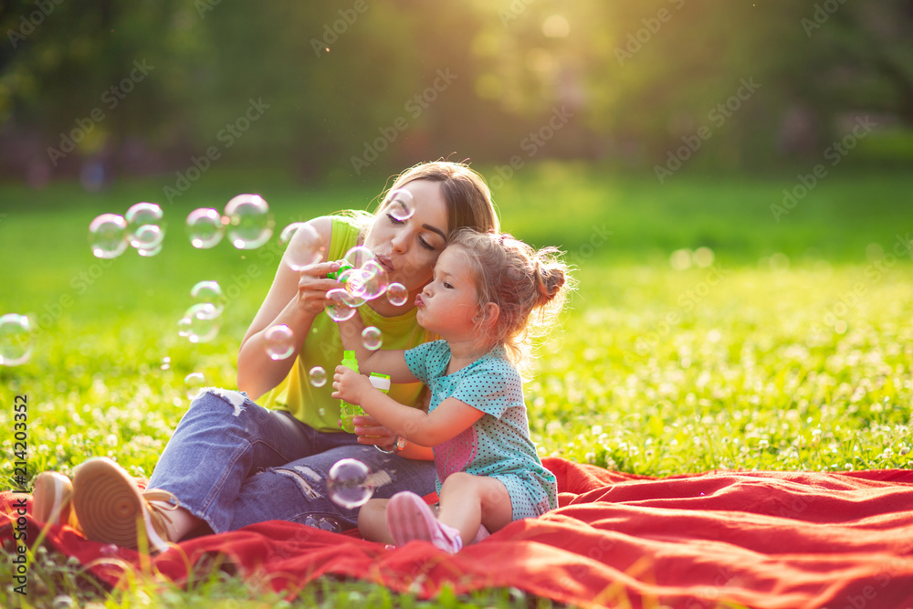Family in park -Female child blows soup foam and make bubbles with her mother in nature. - obrazy, fototapety, plakaty 