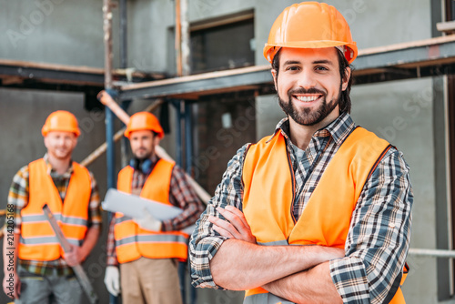 handsome smiling builder standing at construction site with crossed arms while his colleagues standing blurred on background photo