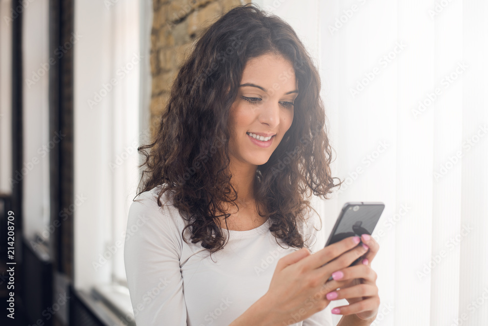 Beautiful girl in casual clothes is using a smart phone and smiling while standing near the window Beautiful girl in casual clothes is using a smart phone and smiling while standing near the window