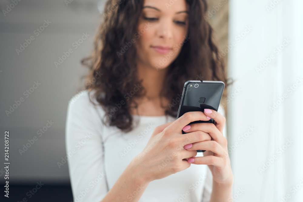 Beautiful curly girl in casual clothes is using a smart phone while standing near the window Beautiful curly girl in casual clothes is using a smart phone while standing near the window