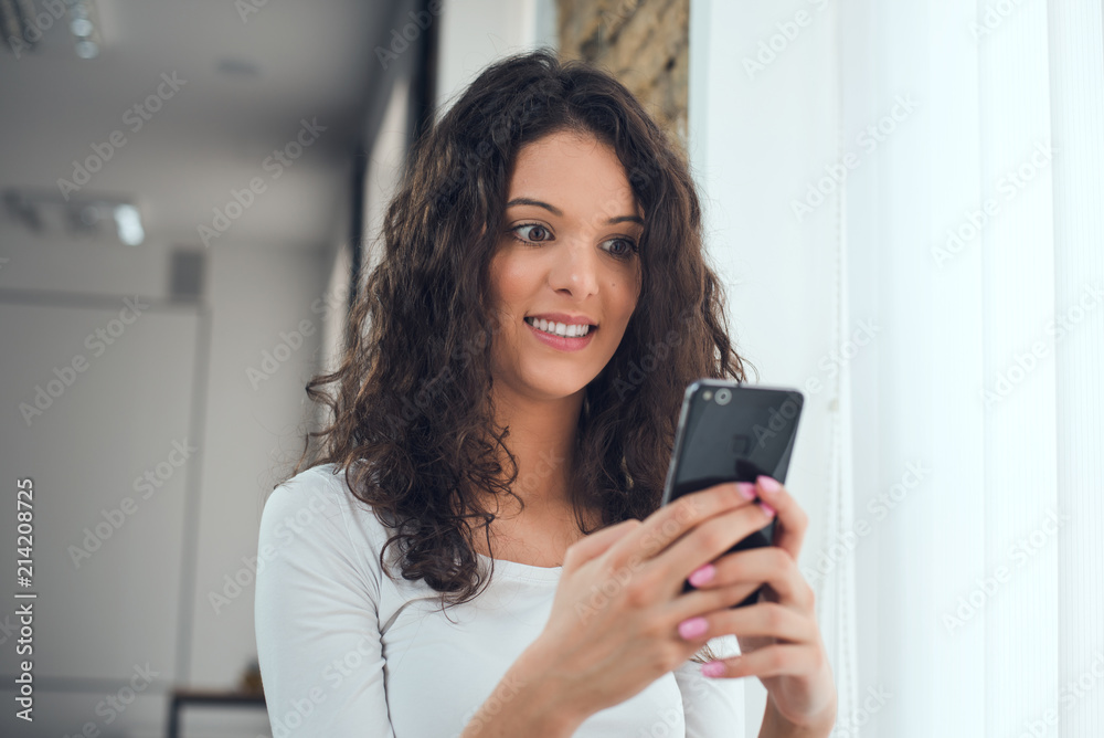 Beautiful curly girl in casual clothes is using a smart phone while standing near the window, surprised face Beautiful curly girl in casual clothes is using a smart phone while standing near the window, surprised face