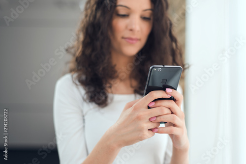 Beautiful curly girl in casual clothes is using a smart phone while standing near the window