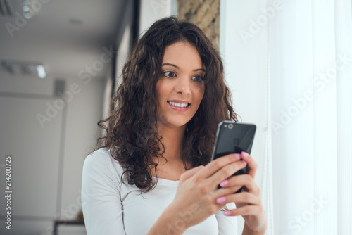 Beautiful curly girl in casual clothes is using a smart phone while standing near the window, surprised face