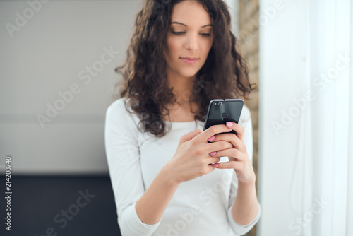 Beautiful curly woman in casual clothes is using a smart phone while standing near the window