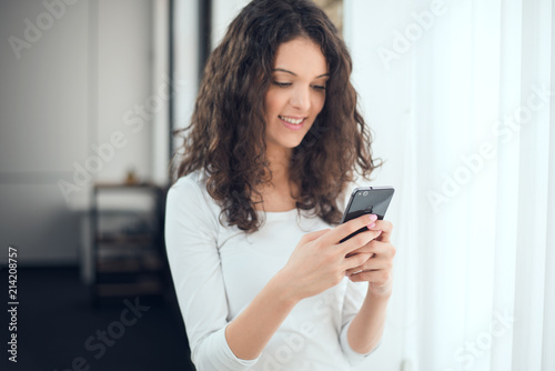 Beautiful woman in casual clothes is texting message and smiling while standing near the window