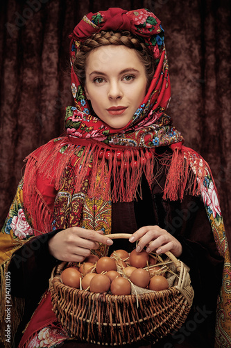 Portrait of  young attractive rural woman sitting with woven basket full of chicken eggs, wearing warm coat, floral shawl and braid wrapped around her head, looking at camera over dark background