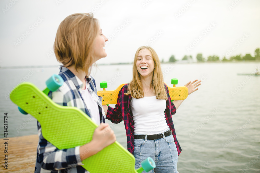 Two cheerful happy skater girls in hipster outfit having fun on a wooden pier during summer vacation Two cheerful happy skater girls in hipster outfit having fun on a wooden pier during summer vacation