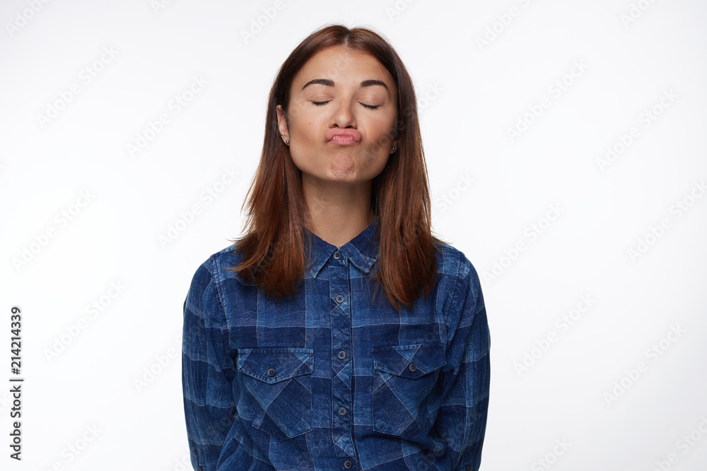 Horizontal portrait of dark haired woman rounds lips as going to kiss boyfriend, closes eyes with pleasure, poses in white studio wall. Cute American young female makes grimace, stands alone in shirt. Horizontal portrait of dark haired woman rounds lips as going to kiss boyfriend, closes eyes with pleasure, poses in white studio wall. Cute American young female makes grimace, stands alone in shirt.