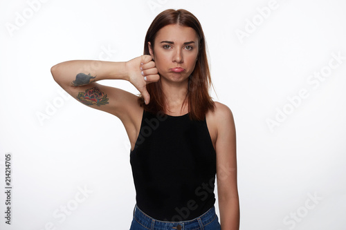 Brunette Caucasian young female expresses dislike with body language, keeps thumb down and frowns face, has displeased face, curves lips, isolated over white studio wall. This is really bad idea!