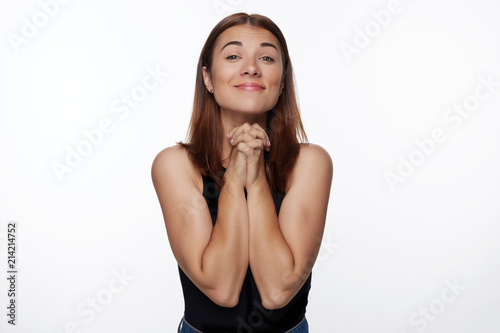 Do favor for me, please! Beautiful brunette female clasps hands together, makes praying gesture and looks with begging expression, wears denim jeans and black top, poses indoor. Body language concept.