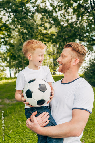 happy father and son holding soccer ball and smiling each other at park