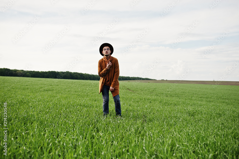 Stylish man in glasses, brown jacket and hat posed on green field. Stylish man in glasses, brown jacket and hat posed on green field.