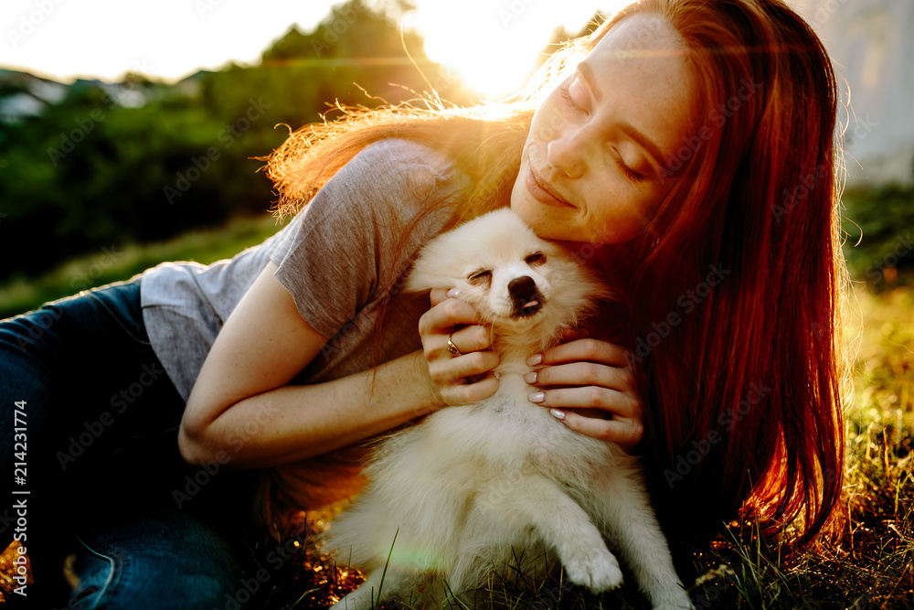 The cute red-haired woman in a hat with a spitz-dog in the park. Beautiful sunset light. Background toning for instagram filter.