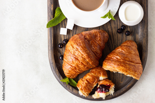 Breakfast concept: two fresh french croissant with a cup of coffee and jam on a dark wooden tray.