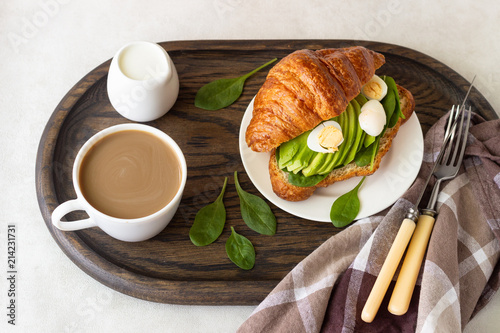 Croissant with green salad, avocado and eggs, a cup of coffee with milk on a dark wooden tray. Breakfast, brunch or lunch concept.