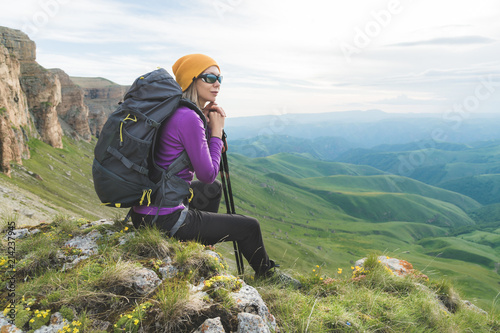 Smiling girl traveler in a yellow hat and a pair of sunglasses ssitting at the foot of epic rocks with a backpack next and looks away photo