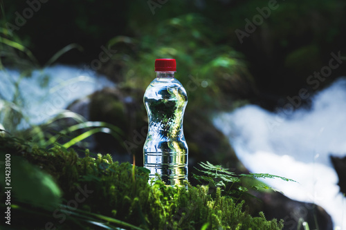 Foto Transparent plastic A bottle of clean water with a red lid stands in the grass and moss on the background of a rugged mountain river