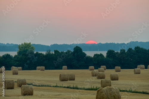 A visible edge of the red sun sits behind a tree in the background of fields with harvested and dried hay.