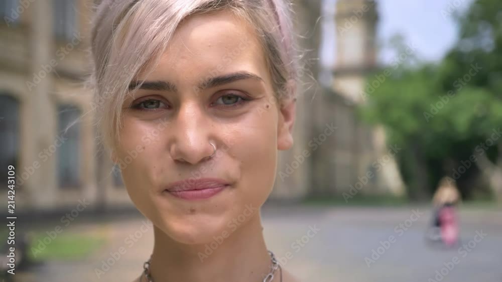 Young blonde woman with pierced nose and short hair smiling at camera, standing on urban street Young blonde woman with pierced nose and short hair smiling at camera, standing on urban street