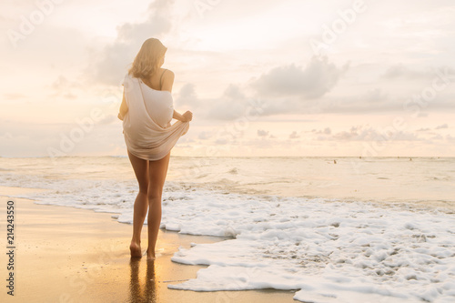 Hipster trendy woman in casual summer dress walk barefoot by the waterline and look to the little waves. Sporty lady on sea sand beach sunset or ocean sunrise. Travel, active, yoga lifestyle concept.