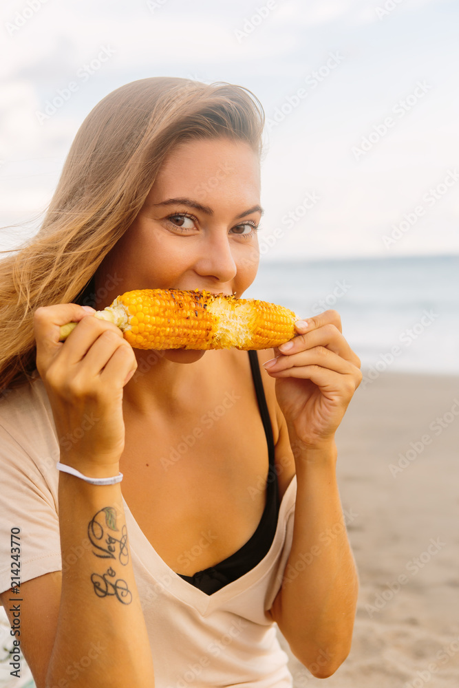 Healthy vegetarian hipster woman in summer outfit eat grilled corn and look to camera. Sexy lady on sea beach sunset or ocean sunrise. Travel, explore, active yoga and meditation lifestyle concept. Healthy vegetarian hipster woman in summer outfit eat grilled corn and look to camera. Sexy lady on sea beach sunset or ocean sunrise. Travel, explore, active yoga and meditation lifestyle concept.