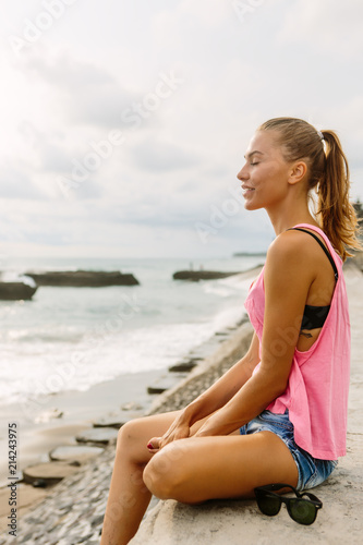 Pretty woman in sport outfit sit on pier and look forward and smile with closed eyes. Sexy lady on sea beach sunset or ocean sunrise. Travel, explore, active yoga and meditation lifestyle concept.
