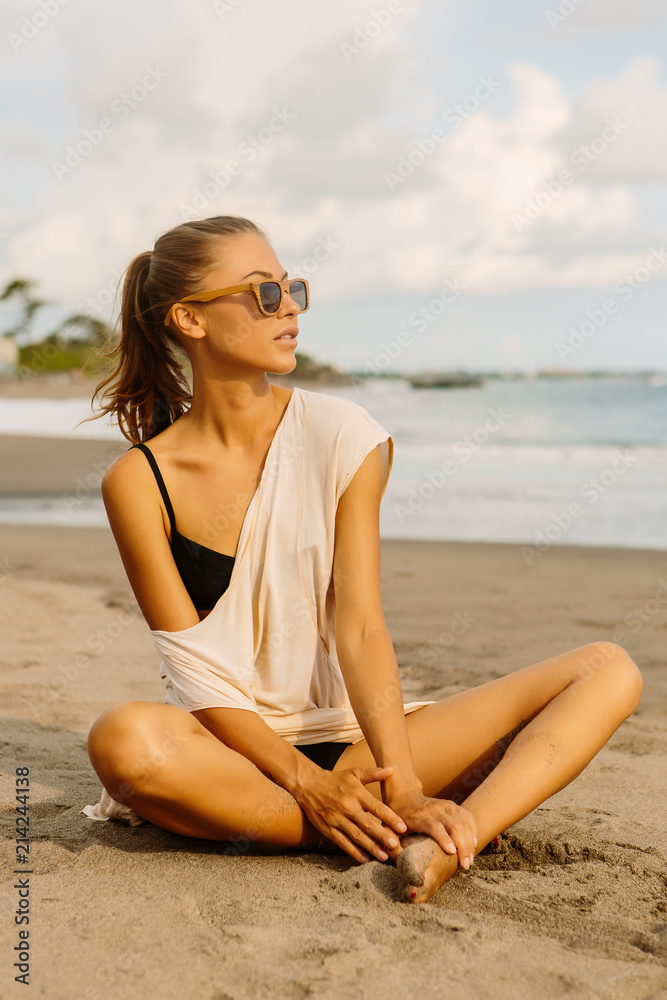 Pretty woman in casual clothes sit on the sand with crossed legs. Sexy lady on tropical exotic sea beach sunset or ocean sunrise. Travel, explore, active yoga and meditation lifestyle concept.
