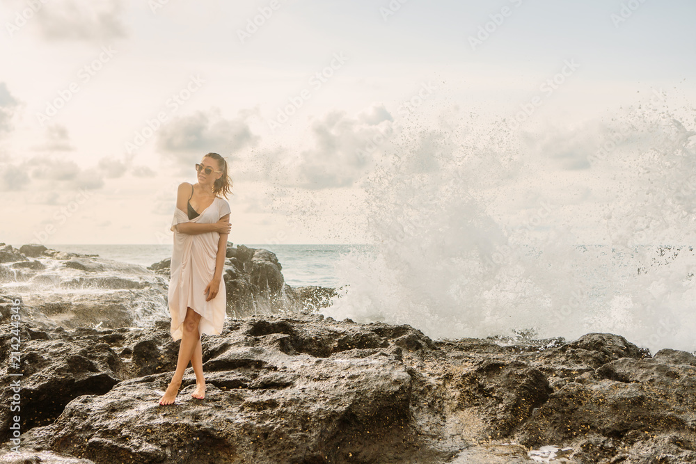 Pretty girl pose to camera in front of huge waves crashing on the stones. Sexy lady on exotic beach seaside sunset or ocean sunrise. Danger, warning, travel, surfer relax and active lifestyle concept. Pretty girl pose to camera in front of huge waves crashing on the stones. Sexy lady on exotic beach seaside sunset or ocean sunrise. Danger, warning, travel, surfer relax and active lifestyle concept.