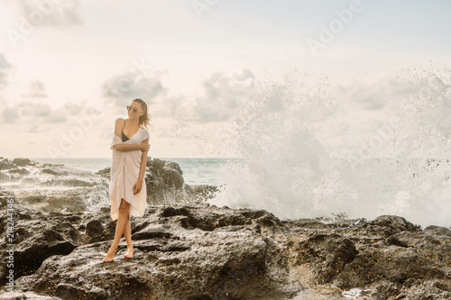 Pretty girl pose to camera in front of huge waves crashing on the stones. Sexy lady on exotic beach seaside sunset or ocean sunrise. Danger  warning  travel  surfer relax and active lifestyle concept.