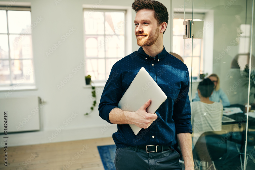 Smiling young businessman walking with a laptop in an office - obrazy, fototapety, plakaty 