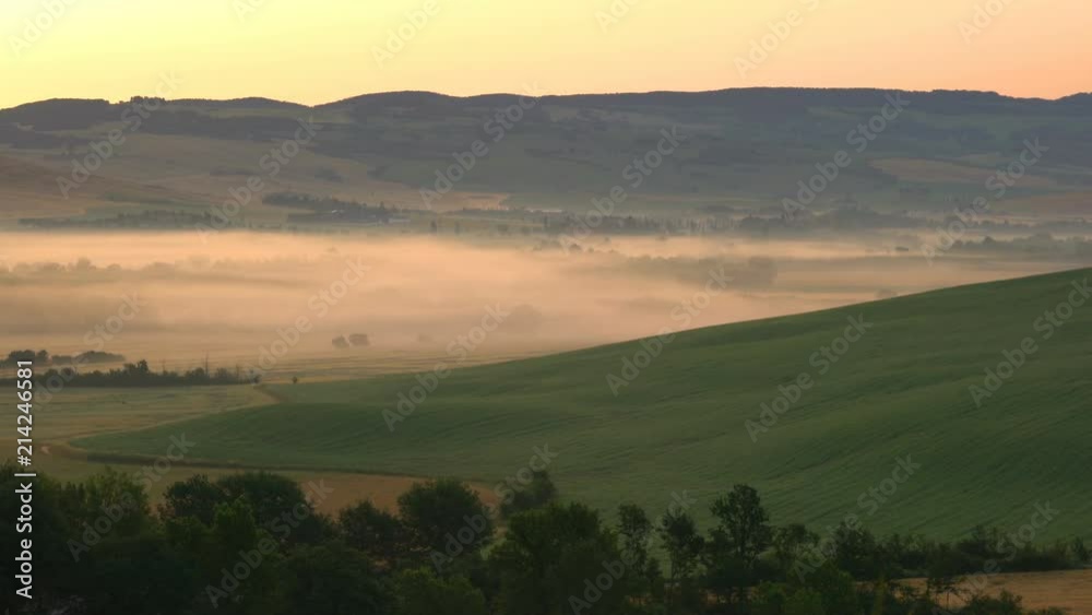 View of beautiful hilly landscape of green tuscan field with fog, line of Cypresses tree and the building at sunrise