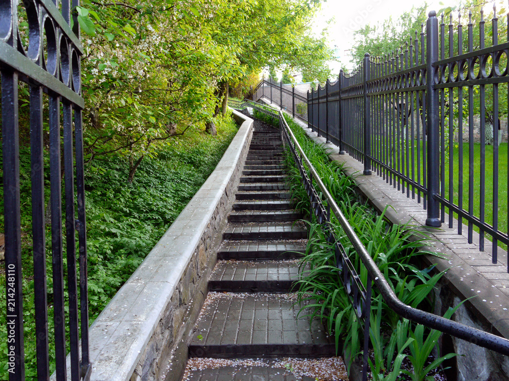 A staircase leading to the top with iron rails. A grating fence on one side and branches of green trees on the other.