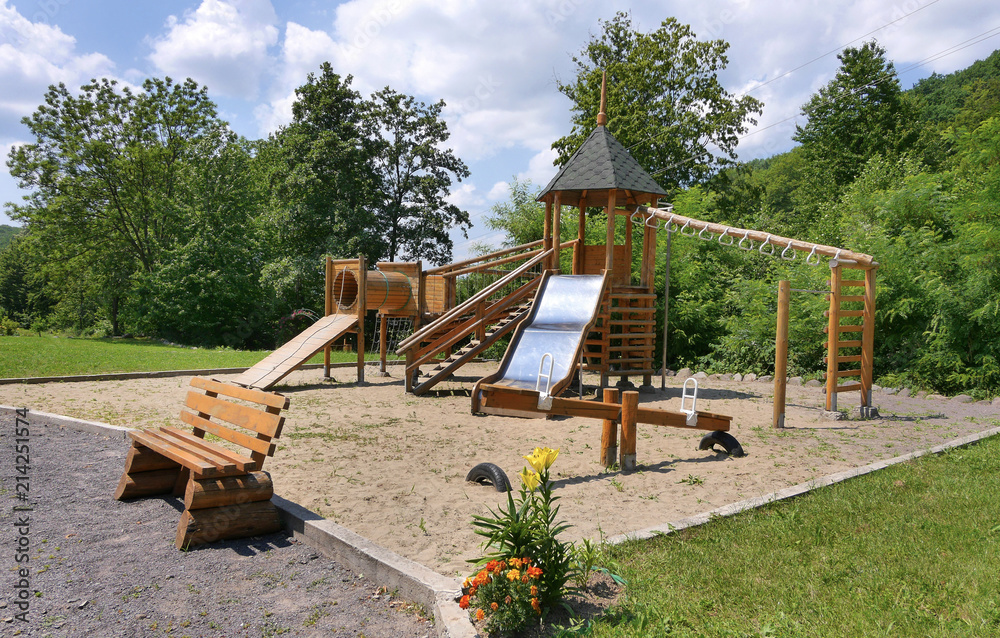 Children's playground covered with sand with a slide for a swing with a wooden deck standing bench for rest and a growing bush color next to it. Children's playground covered with sand with a slide for a swing with a wooden deck standing bench for rest and a growing bush color next to it.