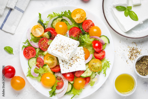 Greek salad with colorful cherry tomatoes red and yellow, cucumber, onion, lettuce and large piece of feta cheese with herbs. In white plate on light background