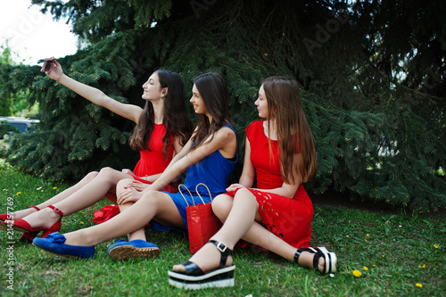 Three teenagers girl in blue and red dresses posed outdoor and making selfie at mobile phone.