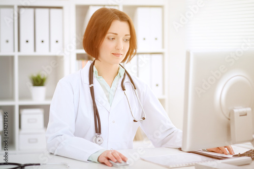 Young brunette female doctor sitting at the table and working  with computer at hospital office
