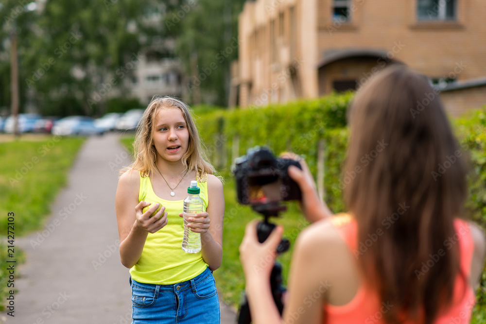 Two girls teenage girlfriends, in city on nature in the summer. In her hands holds a bottle of water and a green apple. Record video on the camera. Concepts of healthy eating. Two girls teenage girlfriends, in city on nature in the summer. In her hands holds a bottle of water and a green apple. Record video on the camera. Concepts of healthy eating.