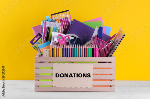 Box with various school and office supplies with a sign on a bright yellow background. Donation concept