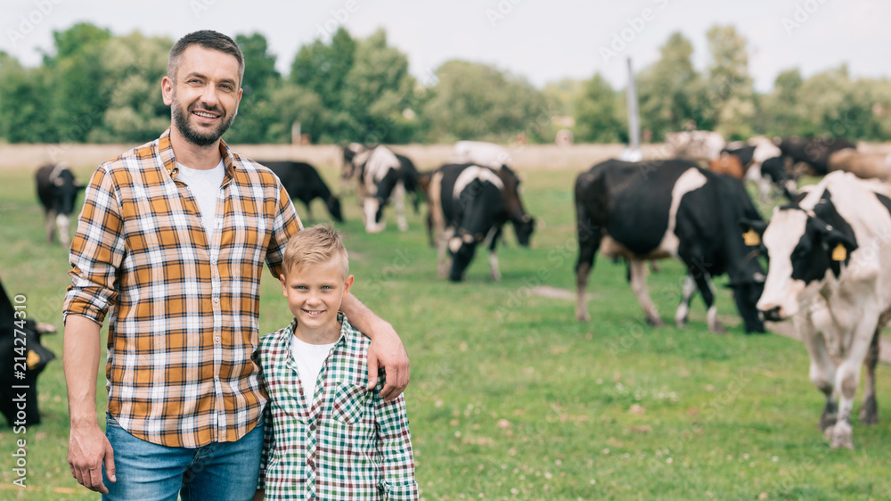 happy father and son smiling at camera while standing near grazing cattle at farm - obrazy, fototapety, plakaty 
