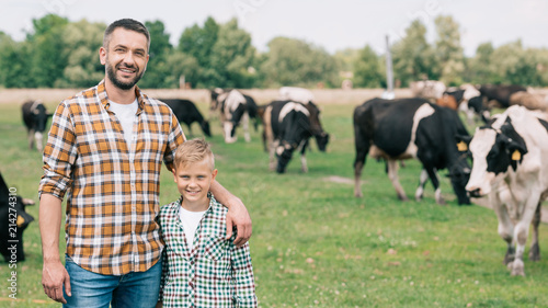 happy father and son smiling at camera while standing near grazing cattle at farm
