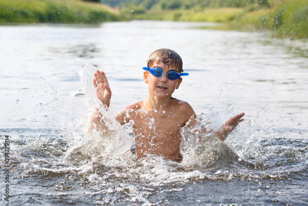 A happy child in swimming goggles splashes and frolics in the water while swimming on a summer day.