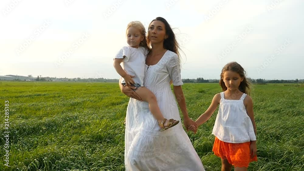 Happy family, mother and her little daughters walk on the green grass, summer evening. Mom with kids walking on green fields. Slow motion Happy family, mother and her little daughters walk on the green grass, summer evening. Mom with kids walking on green fields. Slow motion