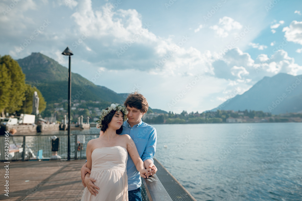 Beautiful young pregnant couple of man and woman standing near the lake Como with scenic mountain view. Sunny summer day. Italy