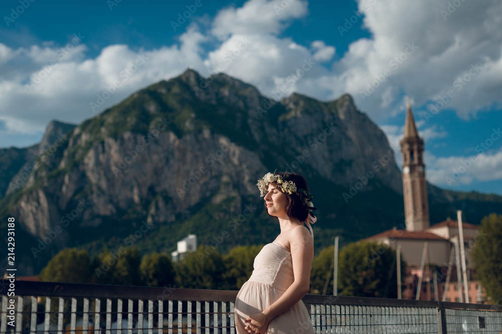 Young tender pregnant woman in creme dress and flower wreath standing near the lake Como with scenic view of rocky mountain. Italy