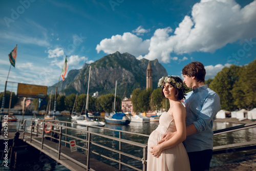 Beautiful young couple of pregnant woman and her husband. Man hugging his wife while standing near the pier on the lake Como, Italy