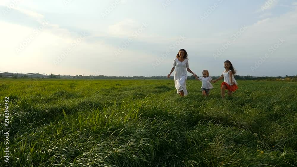 Cute family walking and running on green grassy field. Young mother walks and runs with her little girls, children, on green grass. Slow motion Cute family walking and running on green grassy field. Young mother walks and runs with her little girls, children, on green grass. Slow motion