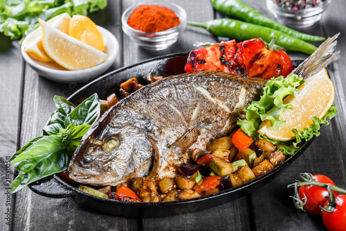 Baked dorado fish with vegetables and lemon on pan on wooden background close up. Delicious dish of seafood