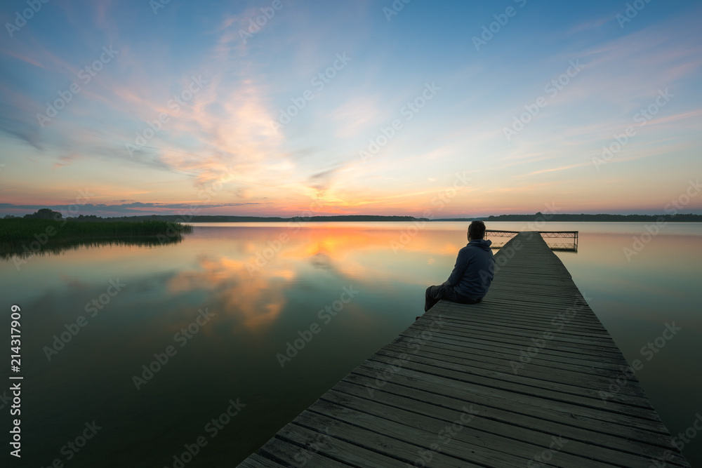 Fototapeta premium Man is sitting on wooden jetty on lake, during sunrise.
