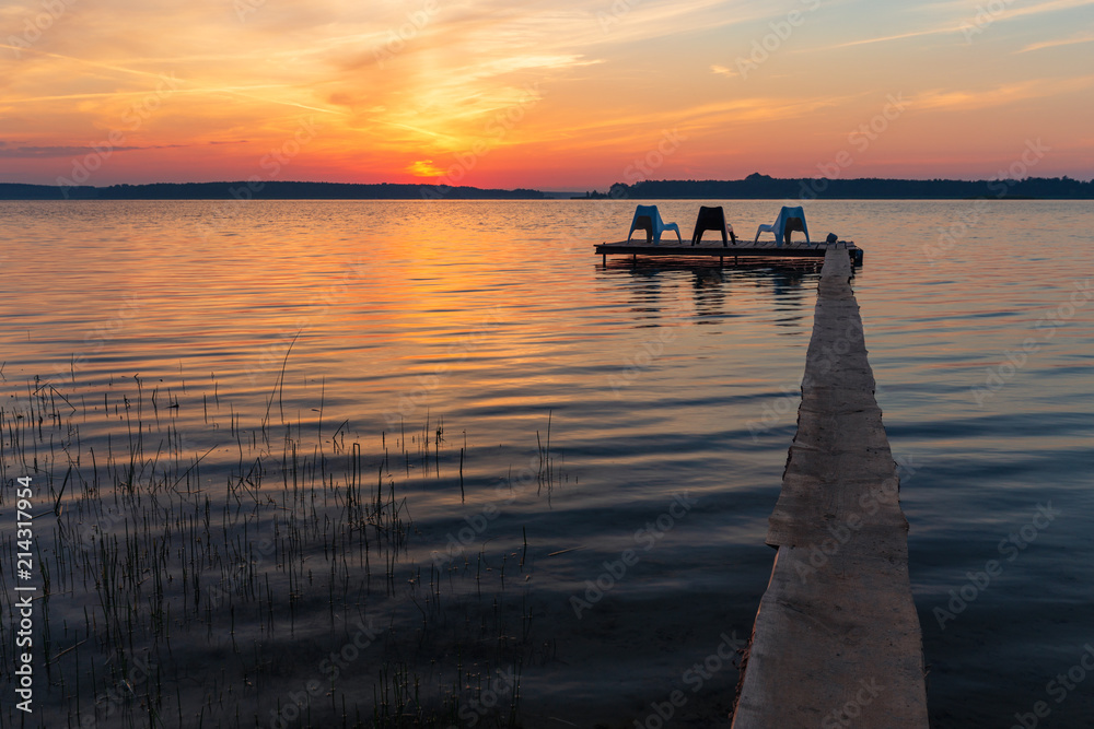 Three empty chairs on wooden jetty on lake, during sunrise.