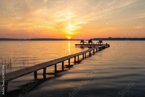 Three empty chairs on wooden jetty on lake, during sunrise.